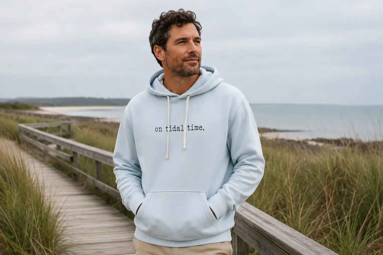 Man wearing a pale blue Great Coast tidal-time hoodie on a coastal boardwalk